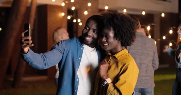 African American young happy and cheerful couple, men and women smiling and posing to smartphone while taking selfie photo at night. late party. Man and woman hugging and having fun. - Footage, Video