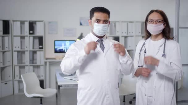 Közepes portré felvétel két modern orvosról, akik fehér köpenyben állnak az irodában, és védőmaszkokat vesznek le, miközben a kamerát nézik. - Felvétel, videó