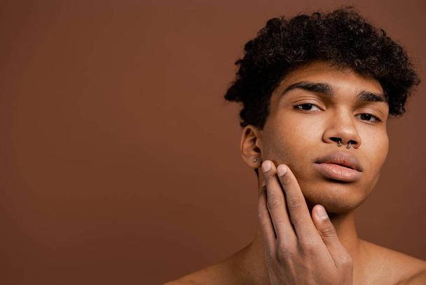 Close up photo of attractive black man with piercing touches his smooth face. Naked torso, isolated brown color background - Photo, Image