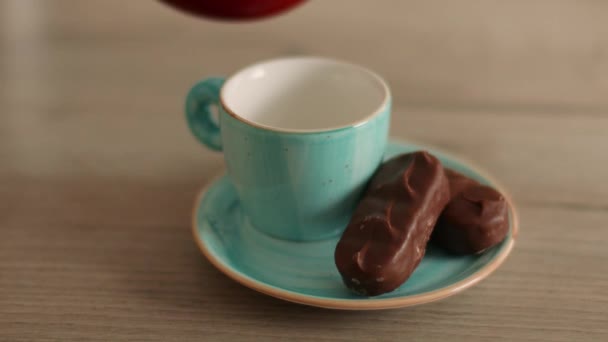 Café negro caliente aromático en taza de cerámica y dulces de chocolate. Verter café negro en taza azul de la cafetera turca en la mesa de madera - Imágenes, Vídeo