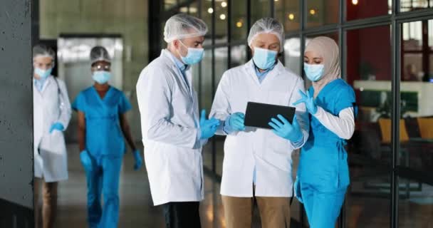 Mixte-races hommes et femmes en hijab médecins en chapeaux, masques médicaux et gants debout à l'hôpital et parler, en utilisant un appareil tablette. Hommes et femmes multiethniques, médecins discutant du diagnostic. Covid-19 - Séquence, vidéo
