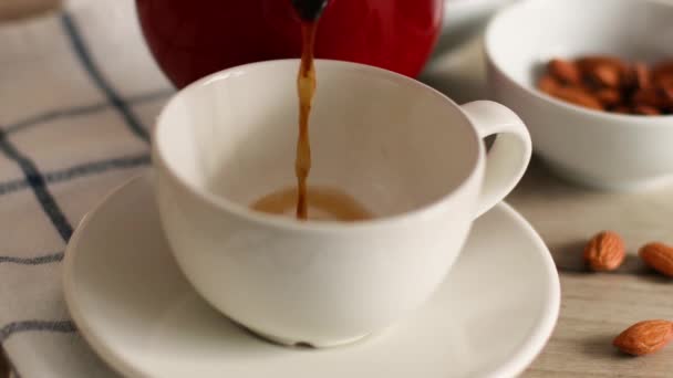Beyaz porselen fincanın içine koyu kahve dökülüyor. Masadaki bir kasede badem fıstığı - Video, Çekim