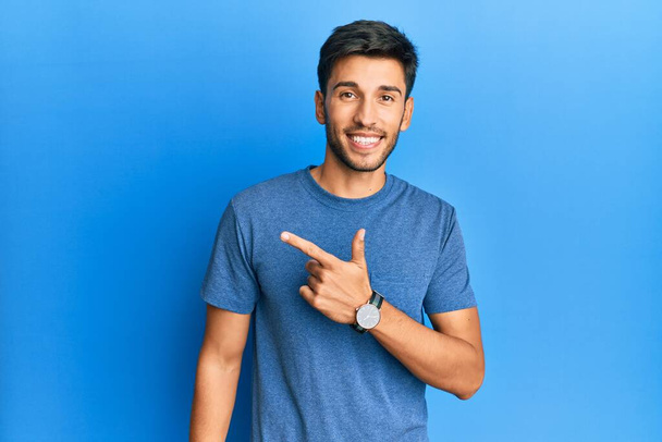 Mavi arka planda sıradan bir tişört giyen genç yakışıklı. Gülümseyen, eli ve parmağı yana doğru olan.  - Fotoğraf, Görsel