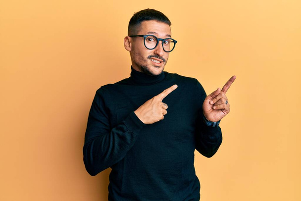 Handsome man with tattoos wearing turtleneck sweater and glasses pointing aside worried and nervous with both hands, concerned and surprised expression  - Photo, Image