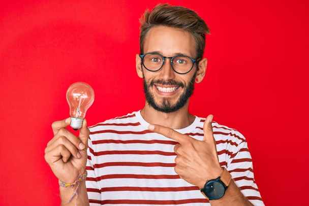 Красивий білий чоловік з бородою тримає лампочку для натхнення та ідеї усміхнений щасливий вказує рукою та пальцем
  - Фото, зображення
