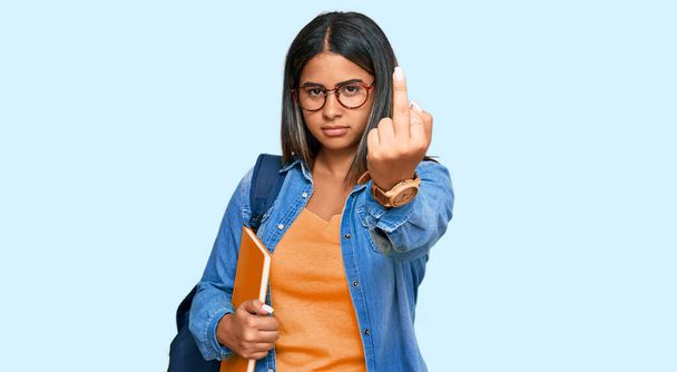 junges lateinisches Mädchen mit Studentenrucksack und Büchern mit Mittelfinger, unhöflichem und unhöflichem Gesichtsausdruck  - Foto, Bild