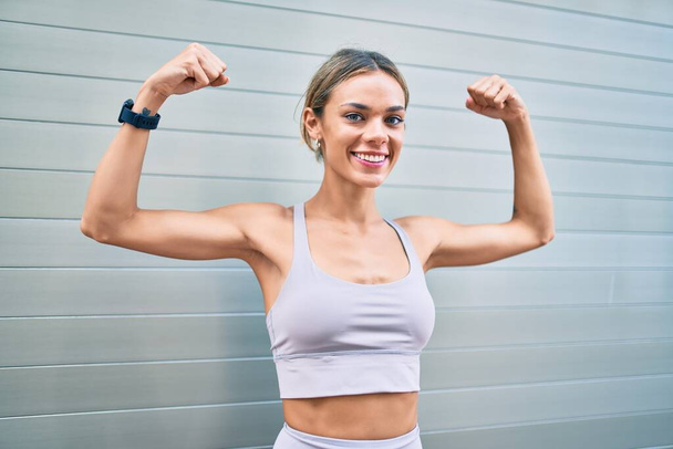 Joven cauciasiano fitness mujer usando deporte ropa entrenamiento al aire libre con los brazos levantados mostrando el músculo orgulloso - Foto, Imagen