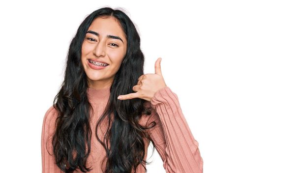 Латиноамериканская девочка-подросток с зубными брекетами в повседневной одежде улыбается, делая телефонный жест с рукой и пальцами, как говорить по телефону. коммуникационные концепции.  - Фото, изображение