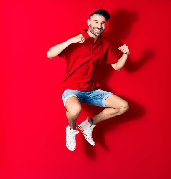 Young handsome deliveryman wearing cap smiling happy. Jumping with smile on face doing winner gesture with fists up over isolated red background - Φωτογραφία, εικόνα