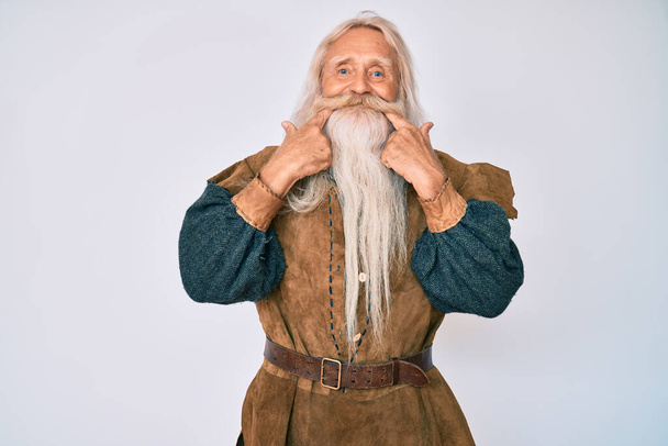 Vieil homme âgé aux cheveux gris et à la barbe longue portant un costume traditionnel viking souriant à la bouche ouverte, les doigts pointant et forçant sourire joyeux  - Photo, image