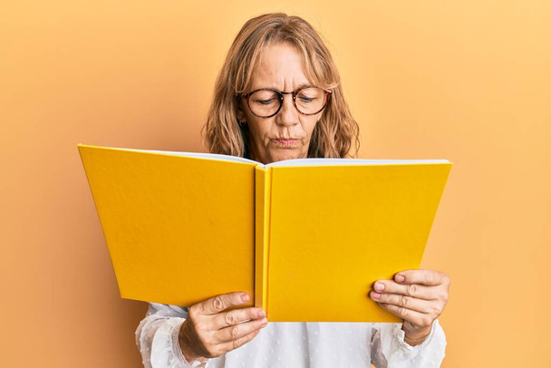 Orta yaşlı sarışın bir kadın kitap okuyor. Gözlüklü, kuşkucu ve gergin. Sorun yüzünden surat asıyor. negatif kişi.  - Fotoğraf, Görsel