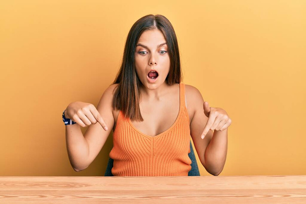 Giovane donna caucasica con indosso abiti casual seduta sul tavolo rivolta verso il basso con le dita mostrando pubblicità, viso sorpreso e bocca aperta  - Foto, immagini