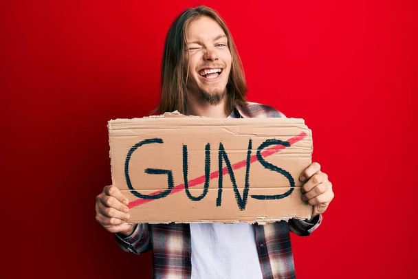 Handsome caucasian man with long hair holding no guns warning banner winking looking at the camera with sexy expression, cheerful and happy face.  - Photo, Image
