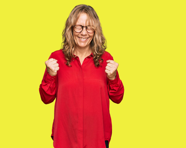 Middle age blonde woman wearing casual shirt over red background excited for success with arms raised and eyes closed celebrating victory smiling. winner concept.  - Photo, Image