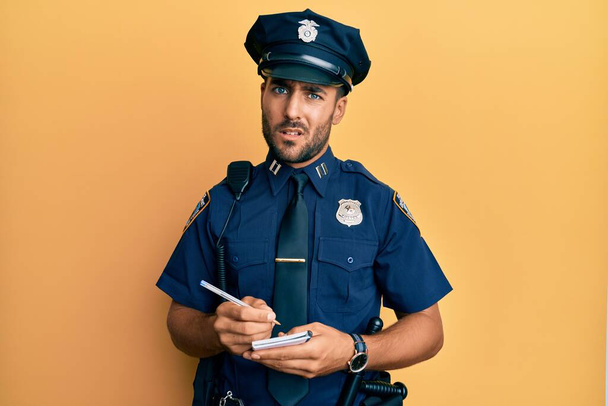 Handsome hispanic man wearing police uniform writing traffic fine clueless and confused expression. doubt concept.  - Фото, изображение