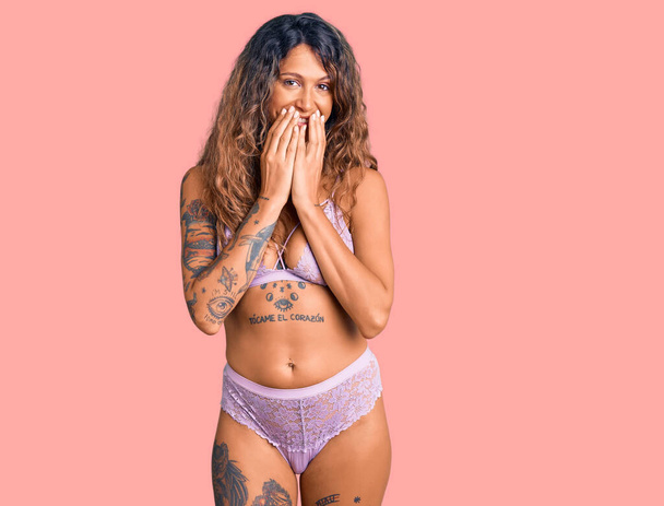 Mulher hispânica jovem com tatuagem vestindo lingerie rindo e riso envergonhado cobrindo boca com as mãos, fofocas e conceito escândalo  - Foto, Imagem