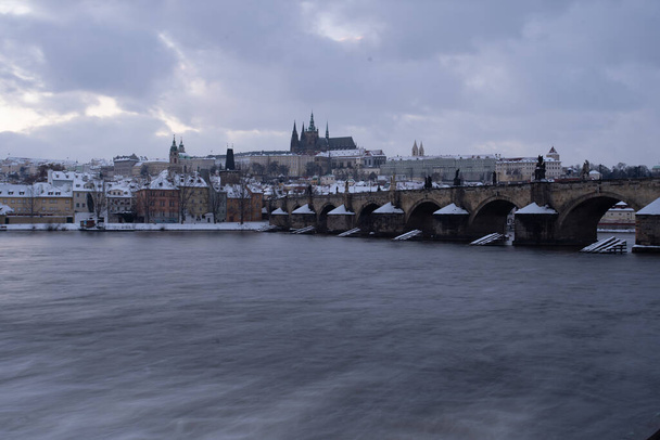Panoramiczny widok na ośnieżony Zamek Praski i katedrę św. Wita oraz Most Karola i jego ośnieżone filary nad Wełtawą wczesnym wieczorem zimy w centrum Pragi - Zdjęcie, obraz