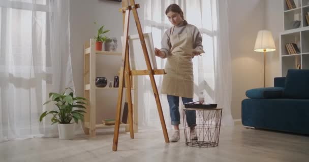 nuori nainen pukeutunut esiliina työskentelee taiteellinen työpaja asunnossa, piirustus kuva ja maalaus väriaineita ja harja kankaalle - Materiaali, video
