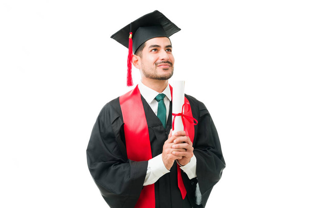 İki eliyle üniversite diploması tutan yakışıklı bir genç. Mezuniyet töreninde gururlu ve minnettar hissediyor. - Fotoğraf, Görsel