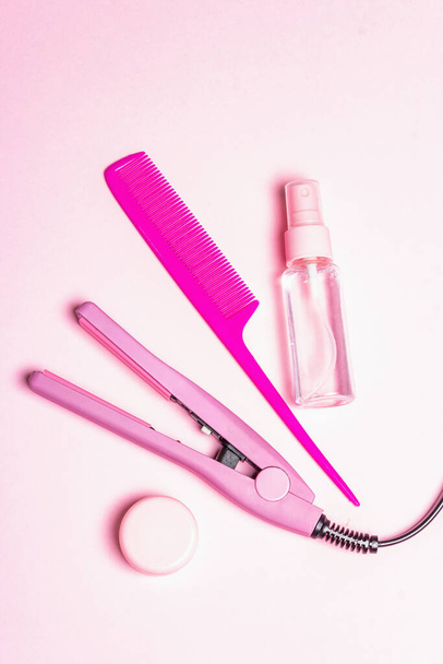 Concept minimaliste de soins capillaires. Un lisseur, une brosse, un spray et de l'huile. Délicat fond rose pastel, vue de dessus - Photo, image