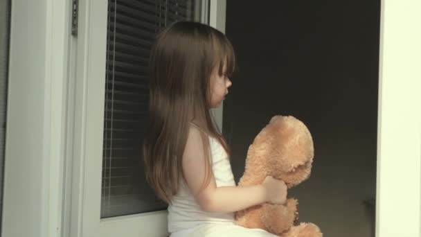 Zorgeloos kind knuffelt zijn speelgoed. Een kind speelt in het voorjaar met een speeltje bij een open raam op vensterbank. Meisje in de zomer speelt met een teddybeer, kijkend uit het raam. Gelukkige jeugd. Dochter speelt - Video