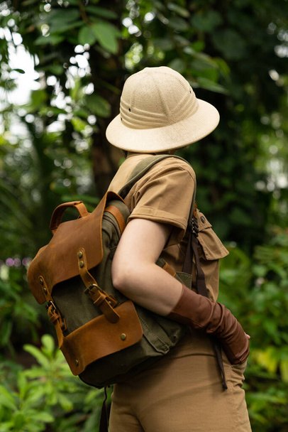 Woman botanist dressed in safari style in greenhouse, back view. Naturalist in khaki clothes, hat, leaver gloves with backpack walks in the rainforest surrounded by palms. Jungle tourist - Photo, Image