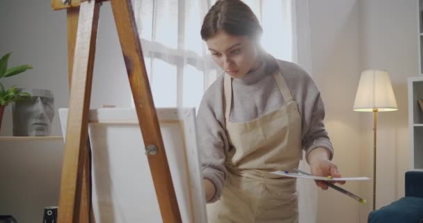 アパートに女性を描く、週末に創造的な趣味、女性アーティストは自宅のスタジオで働いている、イーゼルに絵を描く - 映像、動画