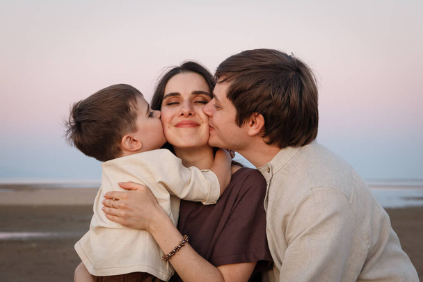 Joven padre con su pequeño hijo besando a su esposa y madre. Ropa de lino de aspecto familiar - Foto, Imagen