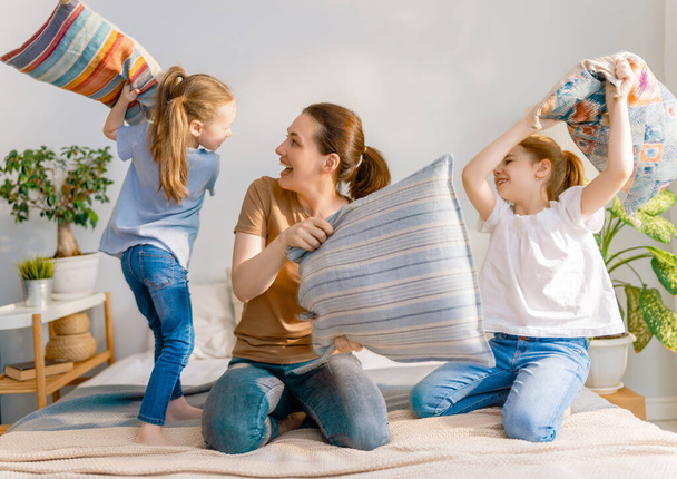 Happy loving family! The mother and her children girls are fighting pillows on bed at home. - Фото, изображение