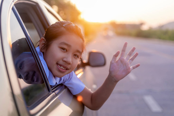 По дороге в школу девочка протянула ему руку из окна машины, смеясь и улыбаясь. Азиатская маленькая девочка улыбается и машет рукой из машины. Дети отдыхают с видом на улицу из машины. Семья в автомобиле. - Фото, изображение