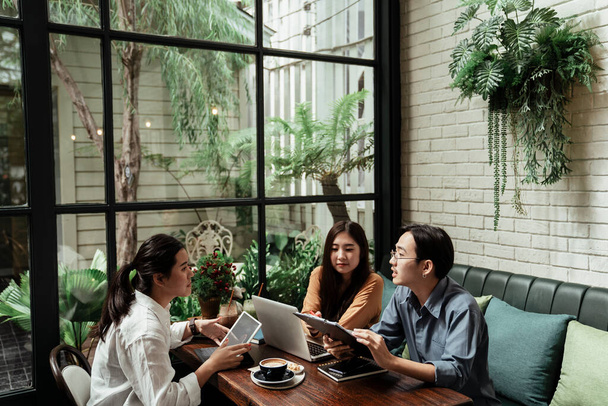 Young startup business people discussing about the plan before invest in something. Young business people talking about the idea of project to their work mate in cafe. Business people metting corporate communication. Three young asian business workin - Photo, Image