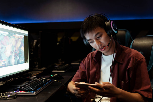 Azjatycki gracz gra na smartfonie, noszenie słuchawki, siedzi w kafejce internetowej. - Zdjęcie, obraz