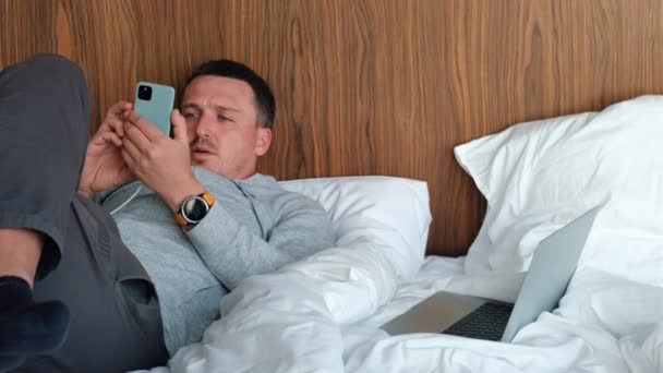 Guy teksty na ładowania smartfona leżącego na łóżku w pokoju hotelowym - Materiał filmowy, wideo