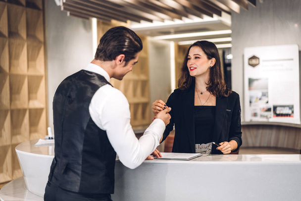 Junge hübsche Geschäftsfrau spricht und checkt Registerinformationen mit einer Empfangsmitarbeiterin ein, die am Schalter der Hotelrezeption in der Lobby eines modernen Hotels steht - Foto, Bild