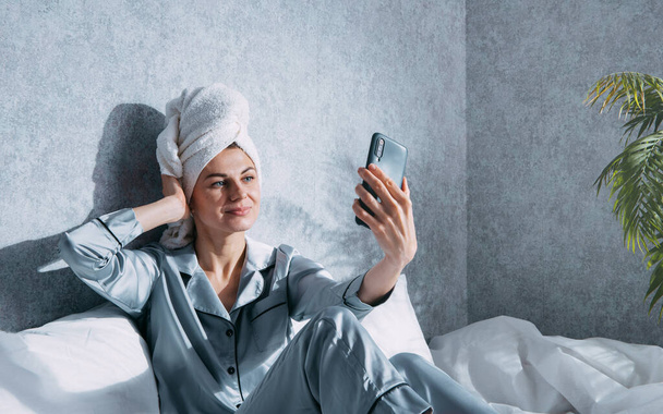 woman in pajamas and a towel on her head sits on a bed in the morning sun and takes a selfie - Zdjęcie, obraz