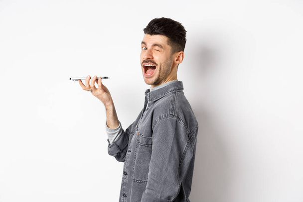 Happy guy winking at looking at camera while using voice translator app or talking on speakerphone, holding phone close to mouth, standing on white background - Photo, Image