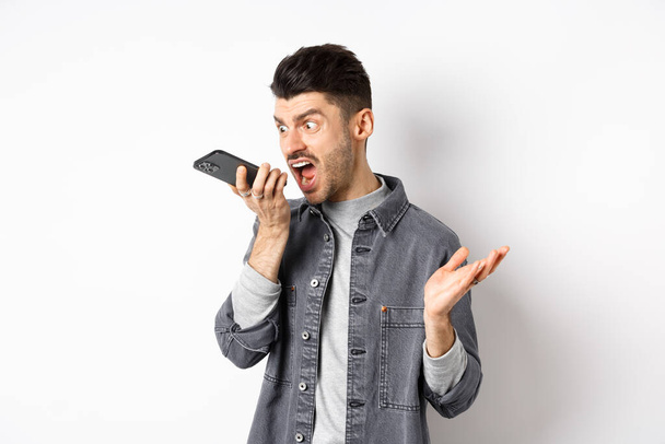 Wütender Typ schreit am Telefon, schreit auf Handy, hinterlässt wütende Sprachnachricht, steht vor weißem Hintergrund - Foto, Bild