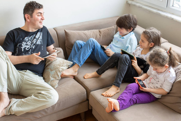 O pai e os filhos estão sentados no sofá. Jogar jogos de internet. todos eles têm celulares e tablets digitais em suas mãos - Foto, Imagem