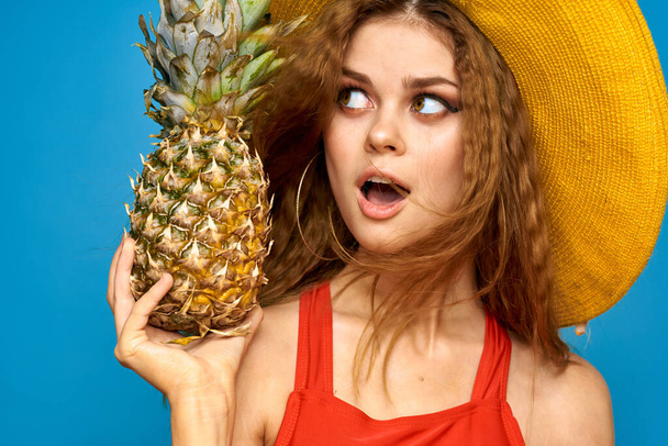 Elinde ananas olan sarı şapkalı bir kadın. Eğlenceli bir yaşam tarzı. Yazın meyve mavisi arka plan. - Fotoğraf, Görsel