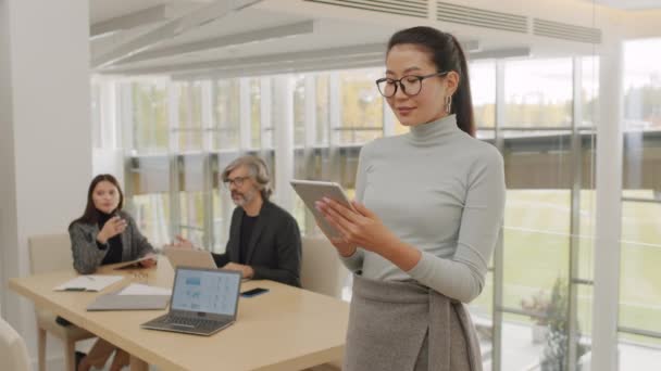 Medium slowmo portret van succesvolle Aziatische zakenvrouw staan met digitale tablet in handen glimlachen om de camera, terwijl haar zakelijke partners onderhandelen op de achtergrond in helder hedendaags kantoor - Video