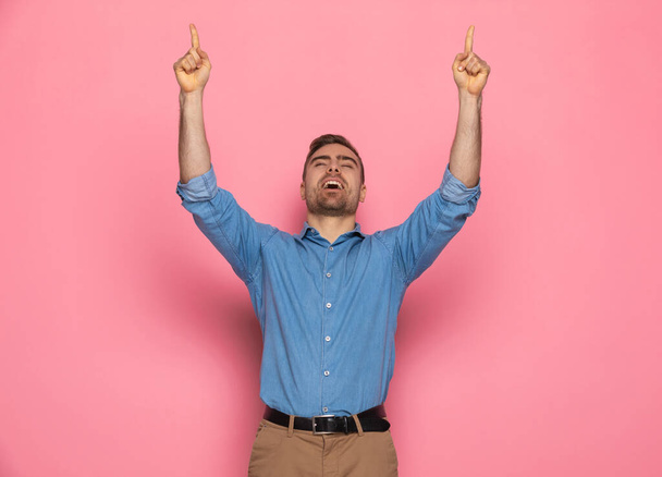 chico joven casual en camisa de mezclilla sosteniendo los brazos por encima de la cabeza, señalando los dedos en el aire y riendo sobre fondo rosa en el estudio - Foto, imagen
