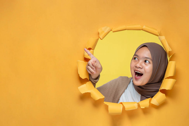 Fröhlich aufgeregter junger muslimischer Teenager aus Asien posiert durch ein zerrissenes gelbes Papierloch und zeigt auf eine Kopierfläche oder leeres gelbes Papier, trägt Hijab, Werbung für das beste Produkt. Werbegeschäft. - Foto, Bild