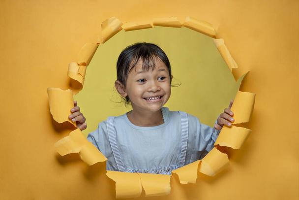 Cute little Asian girl happy smiling poses through torn yellow paper hole, looking something aside. Cheerful little girl - Photo, Image