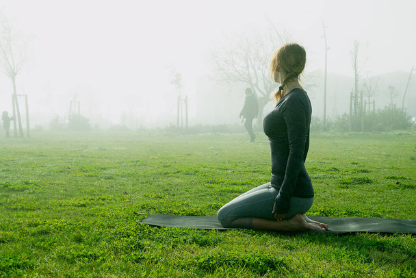Молодая женщина делает yoga, растягивая, exersice, тренировка в парке используя циновку yoga на туманное утро. Природные представления yoga beginner. Концепция здравоохранения. - Фото, изображение