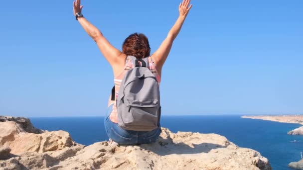 Sırt çantalı genç ve mutlu bir kadın elleri havada bir kayanın üzerinde oturur ve aşağıdaki vadiye bakar. Özgürlük ve kolaylık kavramı. - Video, Çekim