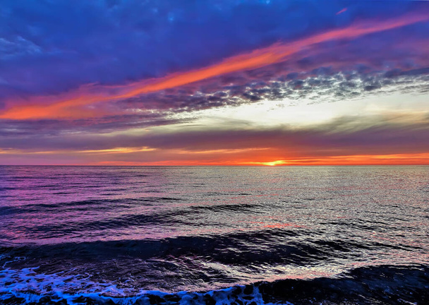 Denizin üzerinde parlak gün batımı. Güneş ufukta kaybolmak üzere. Koyu mavi bulutlar kırmızıyla işaretlenmiş. Su yüzeyinde küçük dalgalar ve köpük var.. - Fotoğraf, Görsel