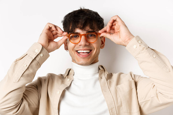 Lähikuva tyylikäs hipster kaveri yrittää silmälasit klo optikko myymälä, laittaa lasit ja hymyillen, seisoo valkoisella taustalla - Valokuva, kuva