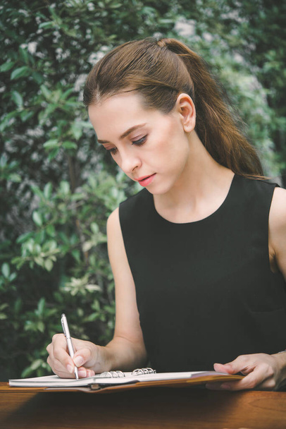 Krásná mladá žena si zapíše poznámku do zápisníku, sedí ráno v zahradě kavárny. - Fotografie, Obrázek