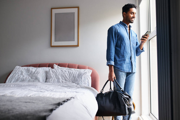 Mies vieras boutique hotellin makuuhuoneessa tarkistamassa sähköposteja ja sosiaalista mediaa matkapuhelimella aamulla - Valokuva, kuva