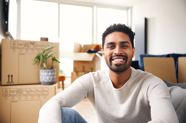 Πορτρέτο του χαρούμενος νεαρός άνδρας που κάθεται στο πάτωμα στο σαλόνι του νέου σπιτιού κοιτάζοντας γύρω καθώς ξεπακετάρει κουτιά απομάκρυνσης - Φωτογραφία, εικόνα